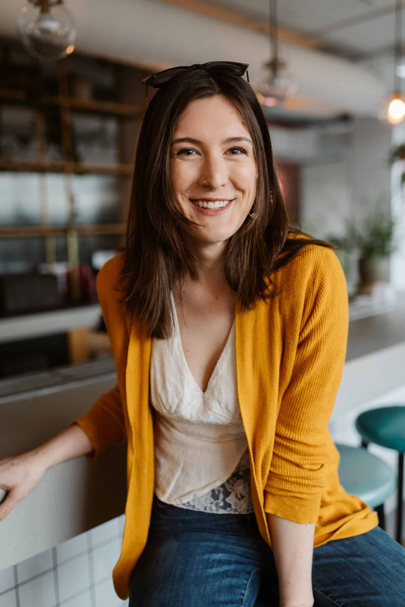 A smiling woman wearing jeans and a yellow cardigan sitting at a bar, showcasing her skills as a WordPress designer.