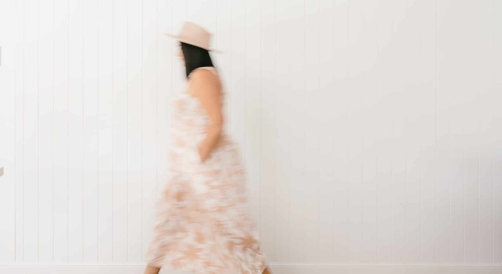 A woman, wearing a hat and dress, walking in a white room.