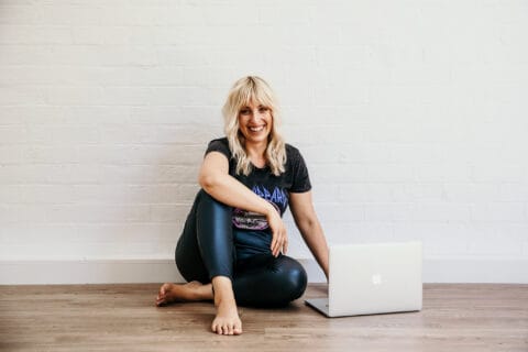 A smiling woman sitting cross-legged on the floor next to an open laptop against a white brick wall background.