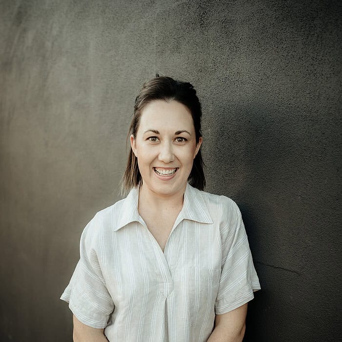 A smiling woman in a white shirt and pants standing against a dark gray wall.