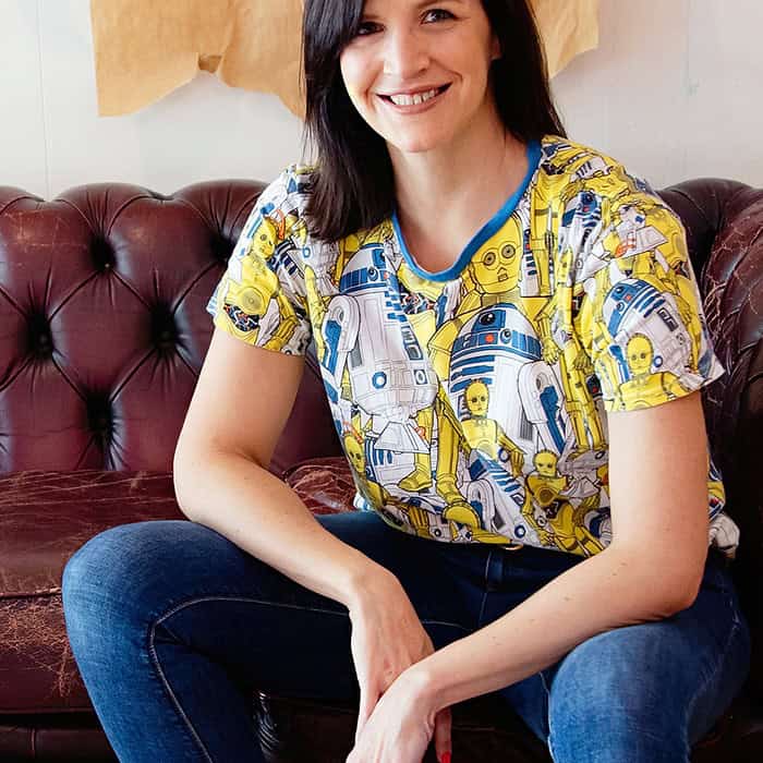 Woman smiling, seated on a brown leather couch, wearing a colorful robot-themed shirt and jeans.
