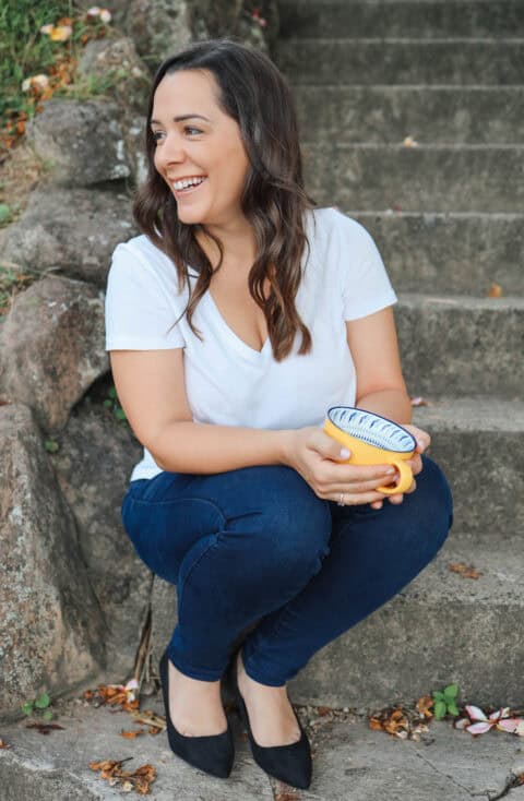 A woman in a white t-shirt and jeans sits on stone steps, holding a banana and a tape measure, smiling off to the side.