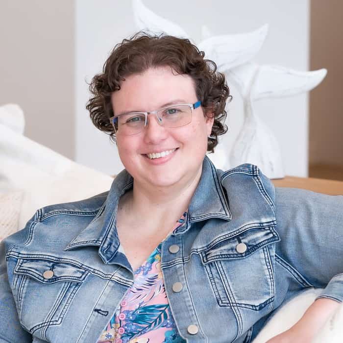 A person with short curly hair, wearing glasses, a denim jacket, and a floral shirt, is sitting on a couch and smiling at the camera.